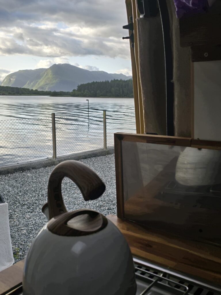 Van Life Europe: Mark, Nikki & Sid’s Journey | Choosing Freedom on the Road 3 Camper van kitchen setup with kettle overlooking lake in Europe