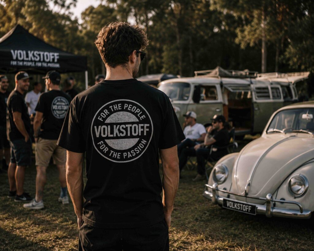 Volkswagen enthusiast wearing a Volkstoff T-shirt at a community gathering surrounded by classic Volkswagens and fellow enthusiasts.