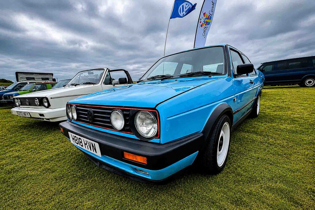 Blue Volkswagen Golf Mk2 GTI displayed on grass at an outdoor VW show, with other classic Volkswagens and club flags in the background.